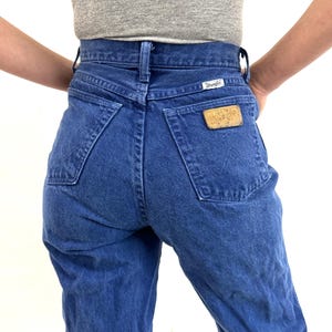 K&ouml;nnte beinhalten: Eine blaue Jeans mit hoher Taille und einem Wrangler-Logo auf der Ges&auml;&szlig;tasche.