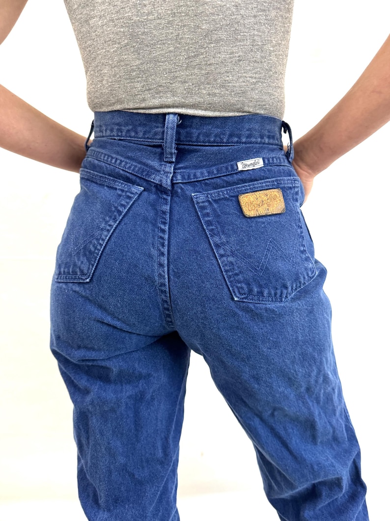 K&ouml;nnte beinhalten: Eine blaue Jeans mit hoher Taille und einem Wrangler-Logo auf der Ges&auml;&szlig;tasche.