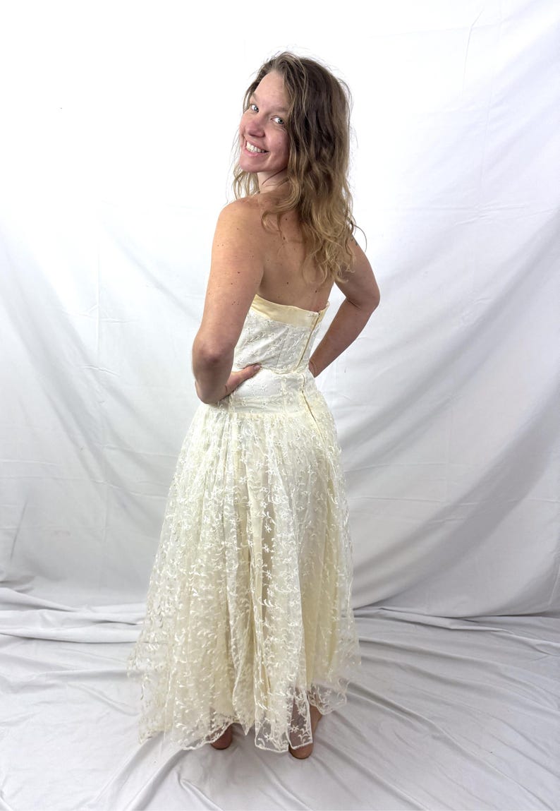 May include: A strapless, ivory-colored dress with a lace overlay. The dress features a fitted bodice and a full, flowing skirt. The model is smiling and looking over their shoulder. The dress appears to be vintage or a formal occasion dress.