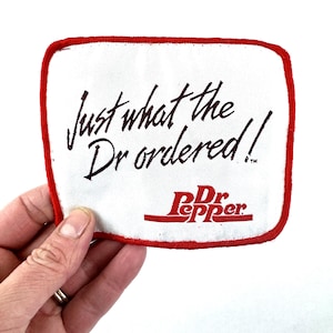 Pode incluir: Um patch branco com uma borda vermelha com o texto "Just what the Dr ordered!" em script marrom escuro. O logotipo da Dr Pepper é exibido em vermelho na parte inferior. O patch é segurado por uma mão.
