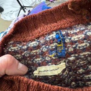 Puede incluir: Primer plano de un su&eacute;ter de lana vintage con cuello acanalado marr&oacute;n y cuerpo estampado en tonos burdeos, azul marino y crema. La etiqueta dice "100% Virgin Shetland".