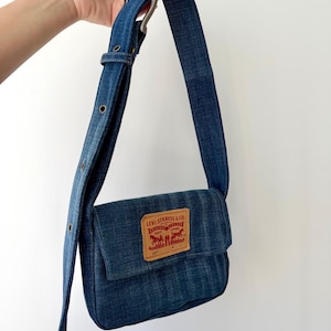 May include: A denim crossbody bag with a flap closure and adjustable strap. The bag features a rectangular leather patch with the Levi Strauss & Co. logo. The strap has a silver-tone buckle and eyelets for adjustment. The bag is held up by a hand.
