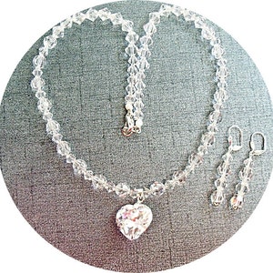 Puede incluir: Un collar de cristal con un colgante en forma de coraz&oacute;n y pendientes de cristal a juego. El collar y los pendientes est&aacute;n hechos de cristales transparentes y plata.