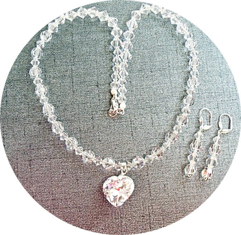 Puede incluir: Un collar de cristal con un colgante en forma de coraz&oacute;n y pendientes de cristal a juego. El collar y los pendientes est&aacute;n hechos de cristales transparentes y plata.