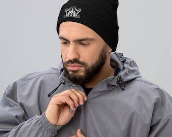Puede incluir: Gorro negro con un diseño bordado blanco en la parte delantera. El diseño bordado presenta una calavera con una corona y otros elementos decorativos. El gorro está hecho de un material suave y lo lleva una persona.