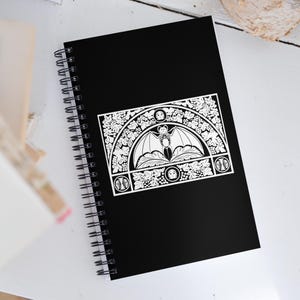 May include: Black spiral-bound notebook featuring a white decorative design of a bat with spread wings, surrounded by floral and geometric patterns. The notebook's cover is solid black, and the design is centered on the front.