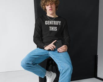 May include: A person wearing a black long-sleeved shirt with the text "GENTRIFY THIS" in white lettering. The person is sitting on a stool and holding a black smartphone.
