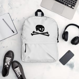 Może przedstawiać: Biały plecak z płótna z czarnym wzorem czaszki i skrzyżowanych kości. Plecak ma przednią kieszeń zapinaną na zamek błyskawiczny i regulowane szelki.