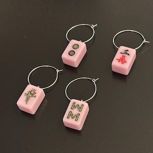 May include: Four pink wine charms with silver hoops. Each charm features a different design from a set of mahjong tiles. The designs include a bird, a circle with a dot, a character, and a double character.