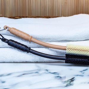 Puede incluir: Dos cepillos para la espalda con mangos largos. Un cepillo tiene un mango marrón y una cabeza de cerdas beige claro. El otro cepillo tiene un mango negro y una cabeza de cerdas negras. Los cepillos están sobre una pila de toallas blancas.