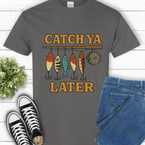 Puede incluir: Camiseta gris con el texto "CATCH YA LATER" en naranja. El diseño presenta una caña de pescar con cinco señuelos de colores colgando. Un par de zapatillas negras y unos vaqueros azules también están en la imagen.