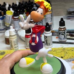 May include: A Rayman figurine, featuring a yellow and orange head, white hands, and a purple body with a red hood. The figure is posed on a green base with white and yellow shoe-like elements. The background includes paint bottles.