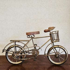 May include: A gold metal bicycle figurine with a brown wooden seat and a wire basket. The bicycle has a chain and gears.