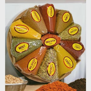 Puede incluir: Una bandeja de madera circular exhibe varios paquetes de especias, como pimentón, comino y orégano. Los paquetes están dispuestos en círculo, cada uno etiquetado con el nombre de la especia. Un cuenco de especias molidas es visible en primer plano.