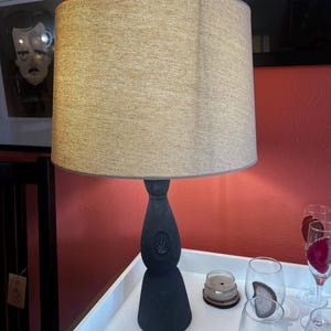 May include: A black ceramic lamp with a beige fabric lampshade sits on a white table. The lamp base is shaped like a bottle with a decorative design. The table is surrounded by glasses with agate coasters.
