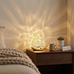 Può includere: Lampada sferica decorativa con superficie strutturata, che emette una luce calda. Si trova su un comodino con una tazza, una piccola pianta in vaso e libri. La luce crea un motivo sulla parete.