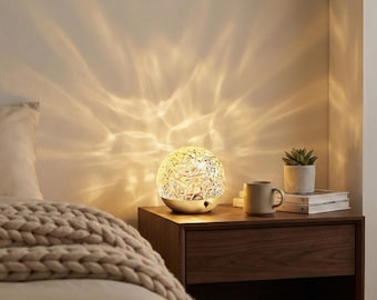 May include: A decorative sphere lamp with a textured, faceted surface, emitting a warm, ambient glow. The lamp sits on a nightstand with a mug, a small potted plant, and books. The light creates a pattern on the wall.