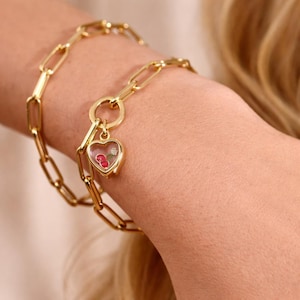 Könnte beinhalten: Goldfarbenes Kettenarmband mit herzförmigem Anhänger. Der Anhänger enthält kleine rote Kirschen und eine weiße Blume. Das Armband besteht aus rechteckigen Gliedern und wird am Handgelenk getragen.