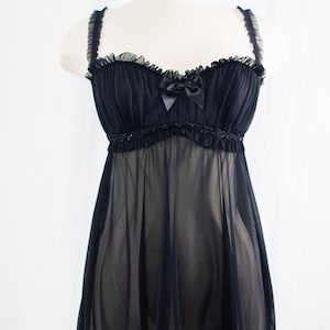 May include: Black babydoll lingerie with a sheer skirt and ruffled trim. The top features a sweetheart neckline, a small bow, and delicate straps. This sleepwear is displayed on a mannequin against a white background.