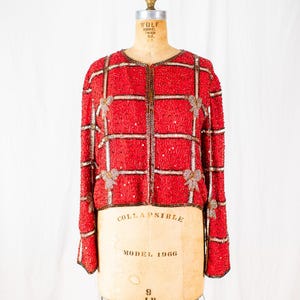 May include: Red beaded jacket with a grid pattern and bow accents. The jacket is displayed on a vintage dress form. The jacket has long sleeves and a round neckline. The dress form is labeled "COLLAPSIBLE MODEL 1966".