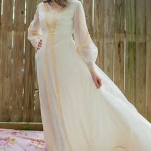 May include: A white vintage wedding dress with long sleeves and gold lace trim. The dress is flowing and has a fitted bodice.