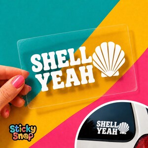 May include: A rectangular clear sticker featuring the white text "SHELL YEAH" in a bold, rounded font next to a white graphic of a seashell. The sticker is shown held up against a colorful background and applied to a vehicle window.