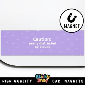 May include: A rectangular car magnet with a light purple background and small white cloud illustrations. The magnet has the text "Caution: easily distracted by clouds" in white. A black and white magnet icon is in the upper right corner.