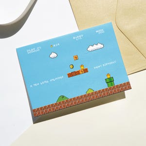 Puede incluir: Una tarjeta de cumplea&ntilde;os azul con un dise&ntilde;o de videojuego pixelado. La tarjeta presenta un nivel con plataformas de ladrillo, tuber&iacute;as verdes y el texto "HAPPY BIRTHDAY!" y "A NEW LEVEL UNLOCKED". La tarjeta va acompa&ntilde;ada de un sobre beige.