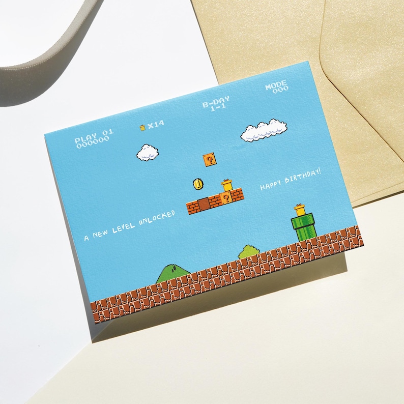 Puede incluir: Una tarjeta de cumplea&ntilde;os azul con un dise&ntilde;o de videojuego pixelado. La tarjeta presenta un nivel con plataformas de ladrillo, tuber&iacute;as verdes y el texto "HAPPY BIRTHDAY!" y "A NEW LEVEL UNLOCKED". La tarjeta va acompa&ntilde;ada de un sobre beige.