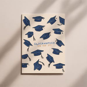 Puede incluir: Tarjeta de graduación con fondo crema y birretes azul marino dispersos por la superficie. El texto "Glückwunsch You Did It!" está escrito en azul. Un artículo de celebración.