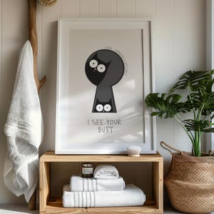 May include: Framed art print featuring a cartoon cat peering through a keyhole shape. The cat is black with large white eyes, and the text "I SEE YOUR BUTT" is below. The print is displayed in a bathroom setting.