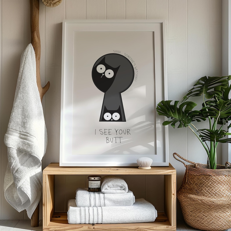 May include: Framed art print featuring a cartoon cat peering through a keyhole shape. The cat is black with large white eyes, and the text "I SEE YOUR BUTT" is below. The print is displayed in a bathroom setting.