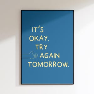 Könnte beinhalten: Gerahmtes Poster mit blauem Hintergrund und den gelben Worten "IT'S OKAY. TRY AGAIN TOMORROW." Eine Hand mit einem Daumen-hoch-Zeichen ist gelb gezeichnet. Der Rahmen ist schwarz.
