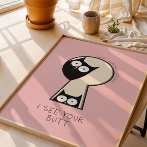 May include: A framed pink art print featuring a whimsical cat design within a keyhole shape. The cat is black and white with large eyes. Text reads "I SEE YOUR BUTT" and "SHHH SOMEONE'S WATCHING!"