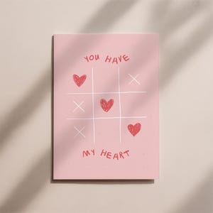 Pode incluir: Um cartão rosa com um design de jogo da velha. O tabuleiro apresenta símbolos de coração vermelhos e X brancos. As palavras "YOU HAVE" estão no topo e "MY HEART" na parte inferior em vermelho.
