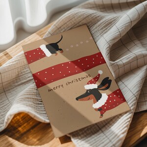 以下が含まれることがあります： ダックスフントのイラストが描かれた長方形のクリスマスカード。茶色の背景に赤い水玉模様のリボンと「merry christmas」の文字が特徴です。ダックスフントはサンタの帽子とコートを着ています。