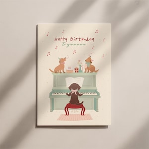 以下が含まれることがあります： 犬のイラストが描かれたバースデーカード。犬がピアノを弾き、他の2匹が歌っています。カードには「Happy Birthday to youuuuu」の文字と音符が描かれています。