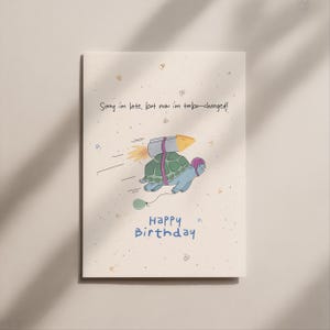 Può includere: Biglietto di compleanno con una tartaruga dei cartoni animati con casco e razzo. Il testo recita: "Scusa il ritardo, ma ora sono turbo-caricato!" e "Buon compleanno" in blu.