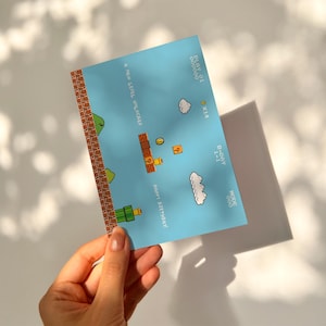 Puede incluir: Tarjeta de cumplea&ntilde;os azul con un dise&ntilde;o de videojuego retro. La tarjeta presenta gr&aacute;ficos pixelados de un nivel de juego, nubes y el texto "A New Level Unlocked" y "Happy Birthday!".
