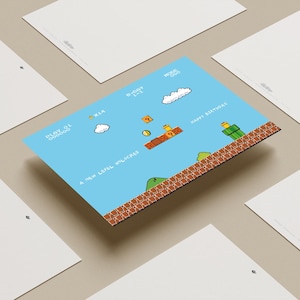 Puede incluir: Una postal azul con un dise&ntilde;o de videojuego pixelado, con el texto "Happy Birthday!" y "A New Level Unlocked." La imagen presenta nubes, monedas y una plataforma de ladrillos. Otras postales blancas rodean la tarjeta principal.