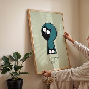 May include: A framed art print with a light green background and a cartoon cat design. The cat is black with white eyes and is inside a teal keyhole shape. The text "I SEE YOUR BUTT" is below the cat. The frame is made of wood.
