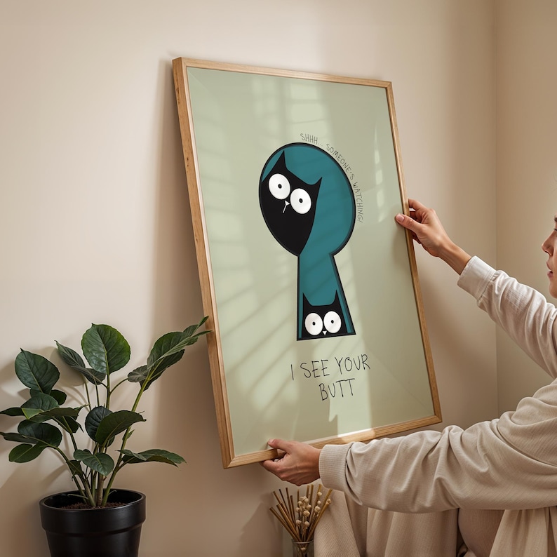 May include: A framed art print with a light green background and a cartoon cat design. The cat is black with white eyes and is inside a teal keyhole shape. The text "I SEE YOUR BUTT" is below the cat. The frame is made of wood.