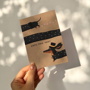 May include: A greeting card with a dachshund illustration. The dog is black with white polka dots and wearing a top hat. The words "Happy New Year!" are printed on the card. The background is tan with a black dotted stripe.