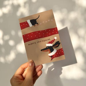 以下が含まれることがあります： サンタの帽子とコートを着たダックスフンドが描かれたクリスマスカード。カードには赤い水玉模様のストライプがあり、茶色の背景に「merry christmas」の文字が書かれています。犬は黒と茶色です。