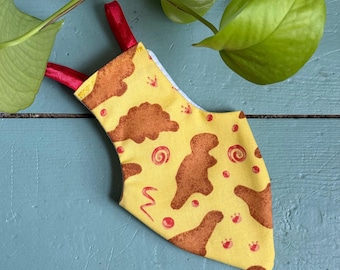 May include: A yellow fabric Christmas stocking with a pattern of brown dinosaur shapes, red swirls, and red dots. The stocking has red ribbon loops for hanging. The background is a blue surface with green leaves.