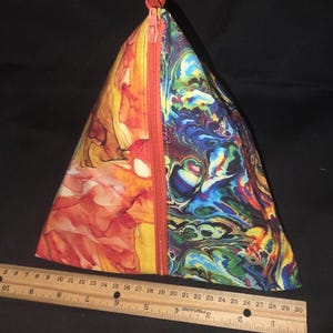 May include: A triangular pouch with a vibrant design. One side features an abstract orange and yellow pattern, while the other showcases a swirling blue, green, and purple design. A red zipper runs down the center. The pouch is approximately 10 inches long.