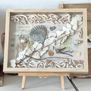 May include: A framed artwork with a collage of seashells, dried plants, and natural elements. It features a large, grey seashell, white coral pieces, and the text "THE OCEAN IS MAGICAL". The frame is made of light-coloured wood.
