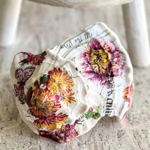 Peut inclure: Coquille d'huître blanche décorative avec un motif floral de fleurs roses, jaunes et rouges, et un papillon. La coquille porte l'inscription "Tell the Story" et d'autres écritures. La coquille est posée sur une surface gris clair.