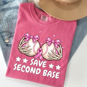 May include: A pink t-shirt featuring a graphic of two baseballs held by skeleton hands, each adorned with a pink ribbon. The text reads "SAVE SECOND BASE" in white letters, accompanied by small white stars and pink hearts. A tan hat and blue jeans are visible.