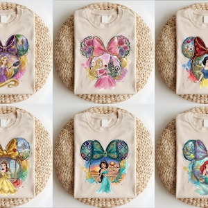 May include: Beige t-shirts featuring a Mickey Mouse head design with a bow and princess illustrations. Each shirt showcases a different princess: Rapunzel, Aurora, Snow White, Belle, Jasmine, and Ariel. The shirts are displayed on woven placemats.